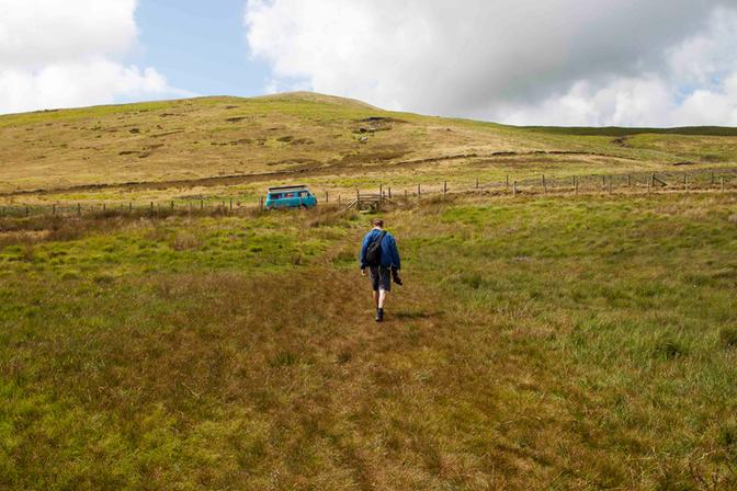 A person walking in a field towards a blue vehicle in the distance, with a hill in the background.