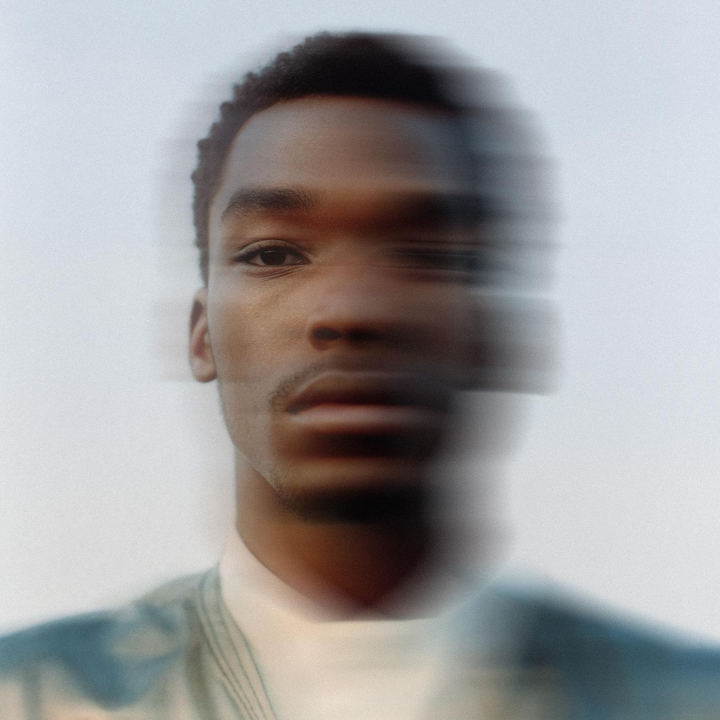 A close-up portrait of a young Black man with a neutral expression, wearing a white shirt and a dark jacket.