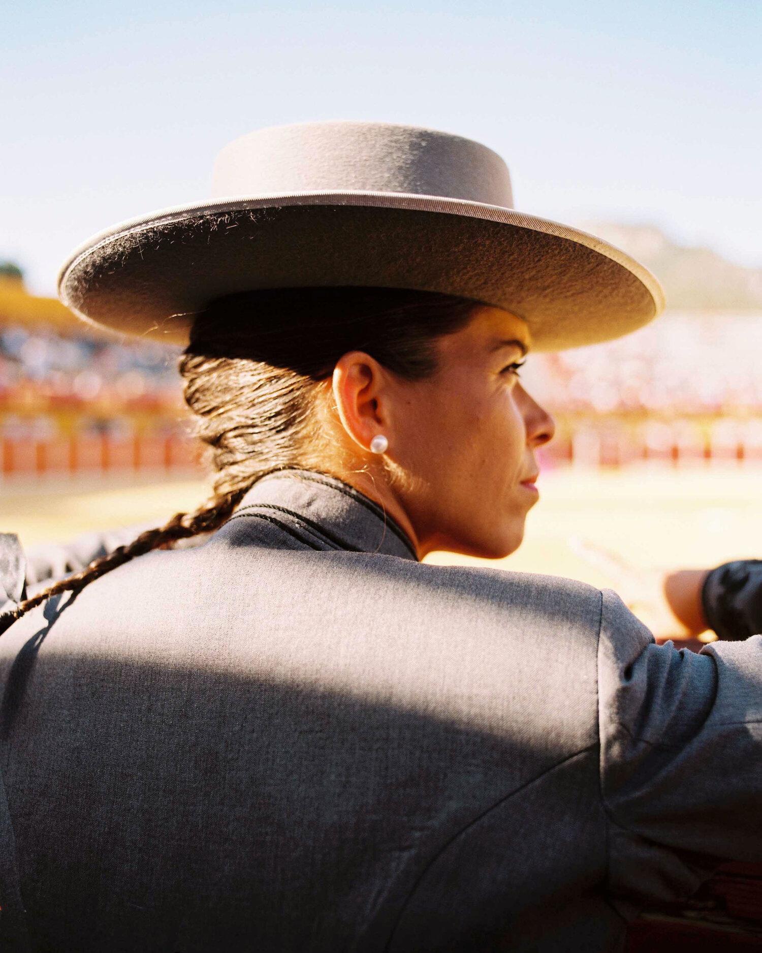 A woman in a traditional Spanish flamenco dress and a wide-brimmed hat, with a long braided hair and a fan in her hand, standing in a crowded outdoor festival setting.