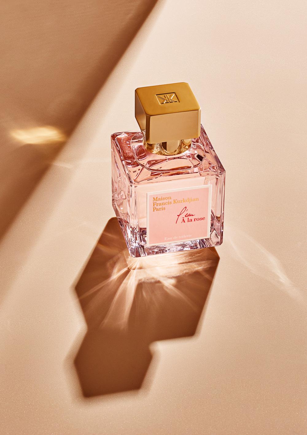 A glass perfume bottle with a gold cap, containing a pink liquid, on a beige surface with a shadow.