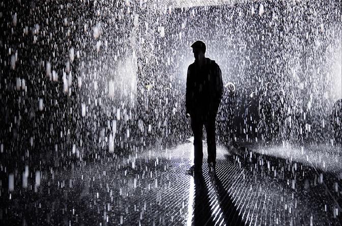A man stands in a room filled with rain, wearing a raincoat and hat.