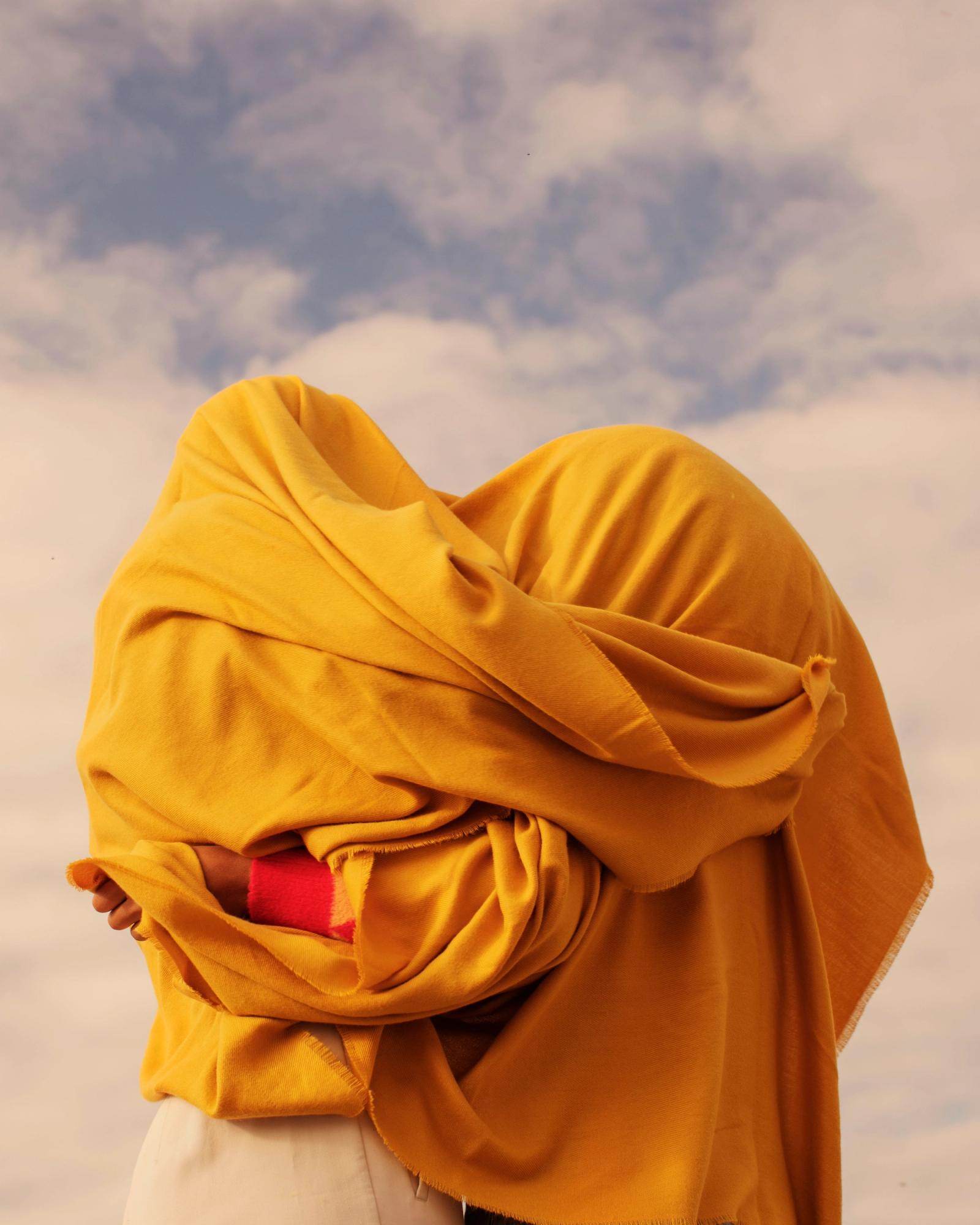 A person wearing a yellow cloak with a red scarf covering their face, standing in a cloudy sky with a few trees in the background.