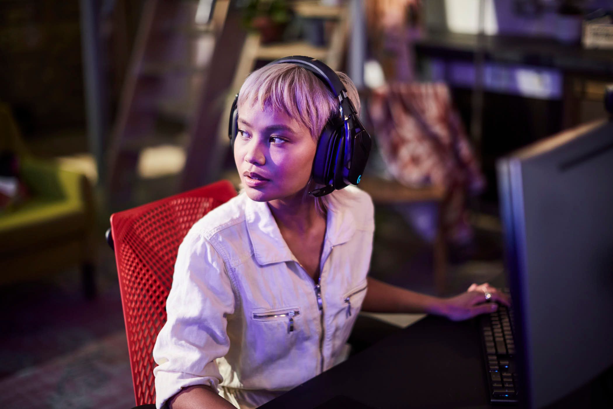 A woman with short blonde hair sits in front of a computer wearing large black headphones. She is wearing a white button-down shirt with a zipper and has a serious expression on.