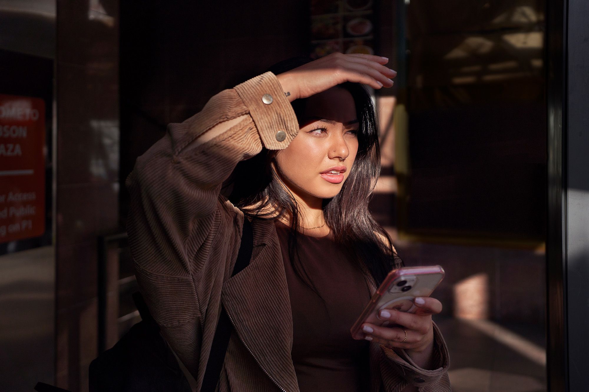 A woman with long dark hair and a brown jacket, looking at her phone with a concerned expression, in a dimly lit urban setting.