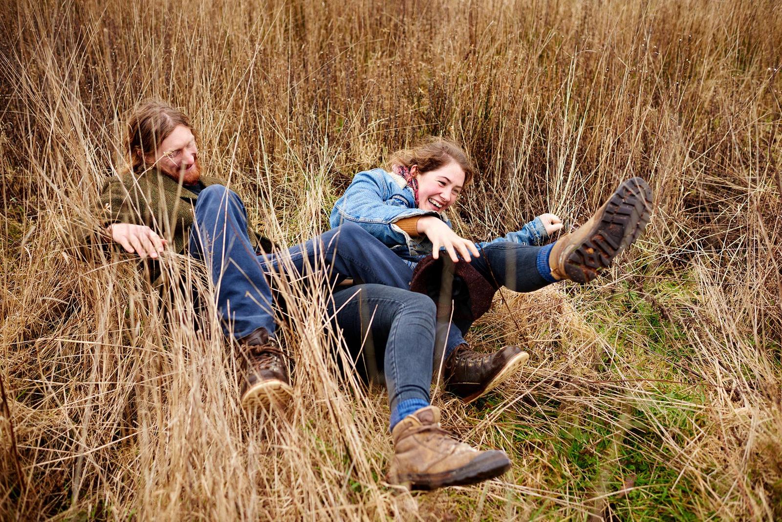 Two people, likely a man and a woman, are sitting in a field of tall, dry grass, laughing and embracing.
