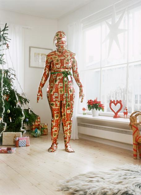 A person is wrapped in a large, red, and white, Christmas-themed, gift-wrapped, paper-like, full-body suit, standing in a room with a Christmas tree, presents, and a window.