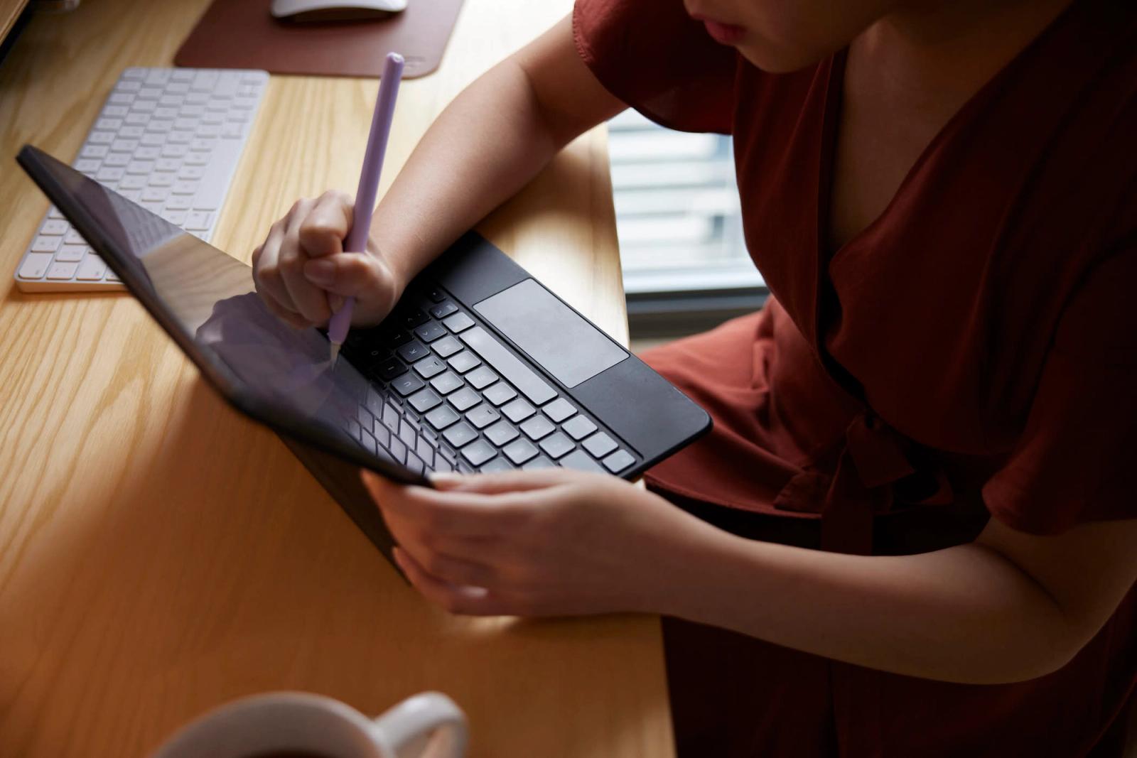 A woman in a red dress is sitting at a desk, working on a laptop. She is holding a pen and looking at the screen with a focused expression.
