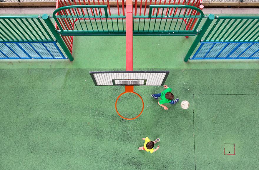 A basketball hoop with a net and a ball on a green playground surface, with a child in a green shirt and blue shorts playing with a ball.