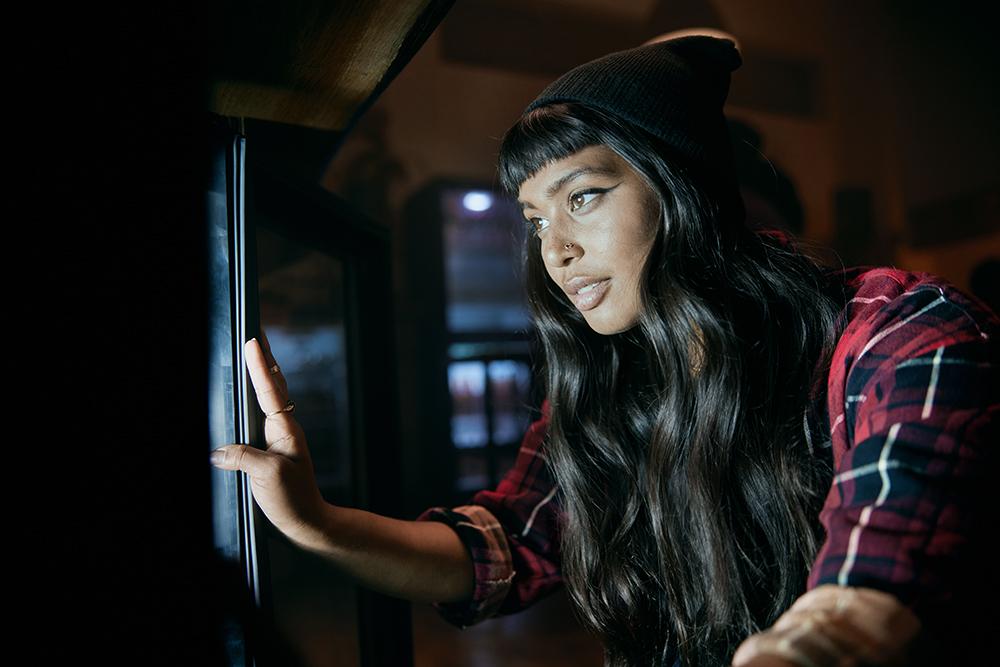 A woman with long dark hair and a red plaid shirt is looking out of a window, wearing a black beanie.