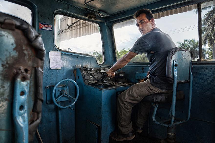 A man in a blue shirt and jeans is sitting in the driver's seat of a train, looking out the window at the viewer.