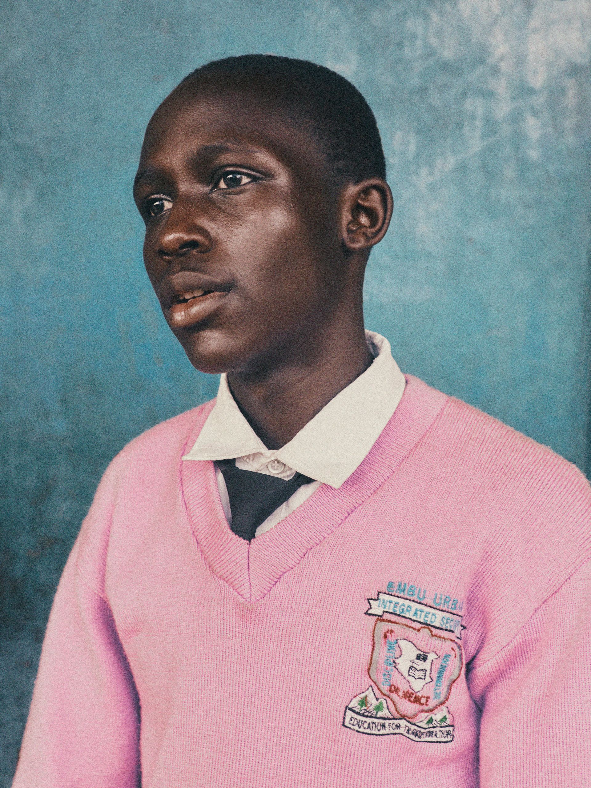 A young boy in a pink school uniform, likely a student at a school in Africa or a similar region, wearing a pink sweater with a school logo on the front.