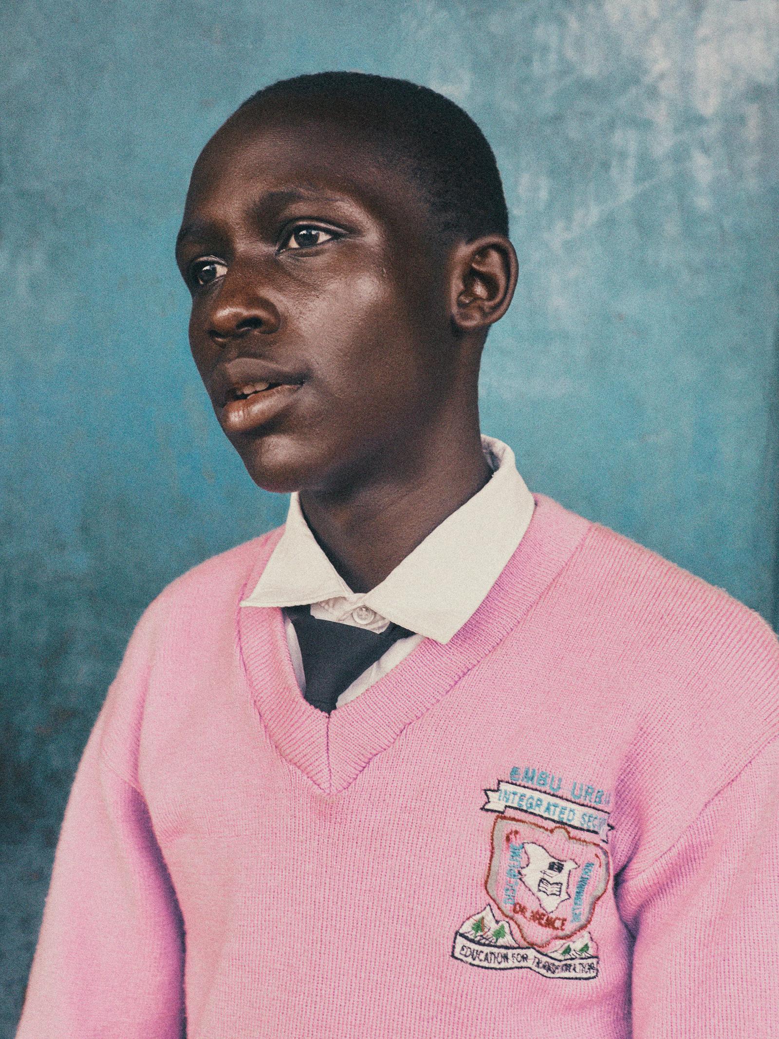 A young boy in a pink school uniform, likely a student at a school in Africa or a similar region, wearing a pink sweater with a school logo on the front.