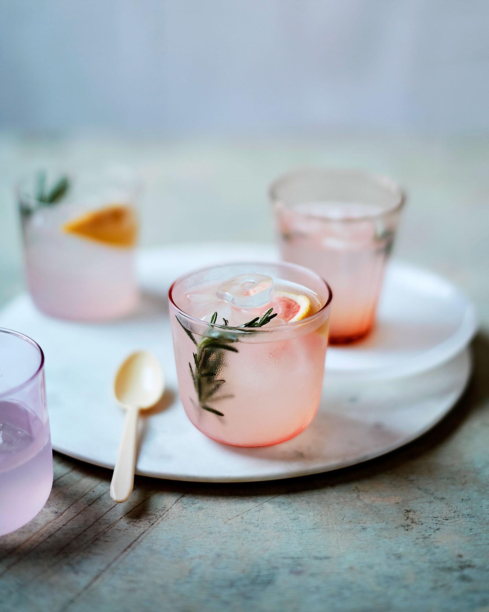 A glass of pink lemonade with a sprig of rosemary and a slice of lemon, served on a white plate with a wooden spoon.