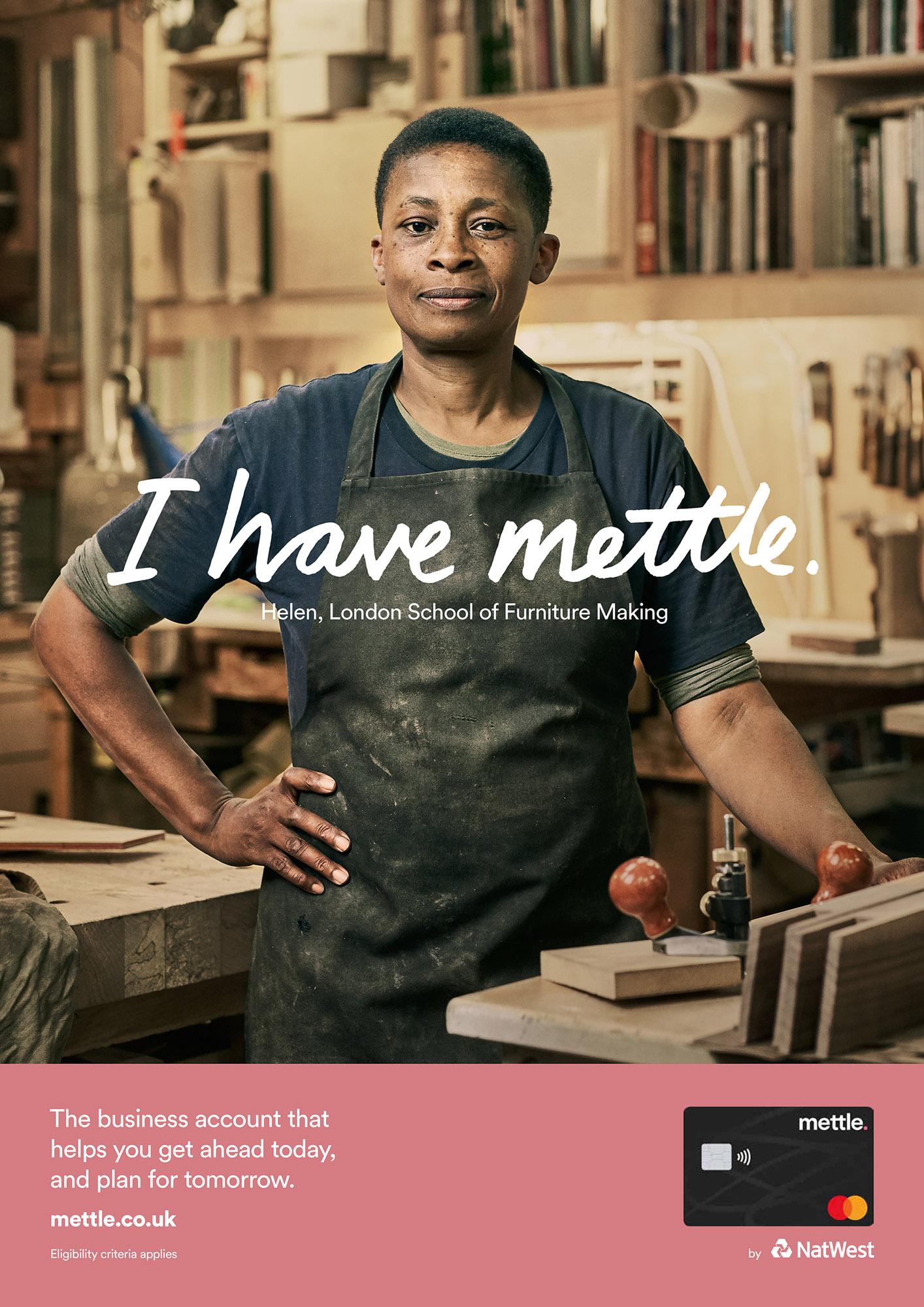 A woman in a black t-shirt and jeans standing in a workshop, holding a piece of wood and a hammer, with a bookshelf and a desk in the background.