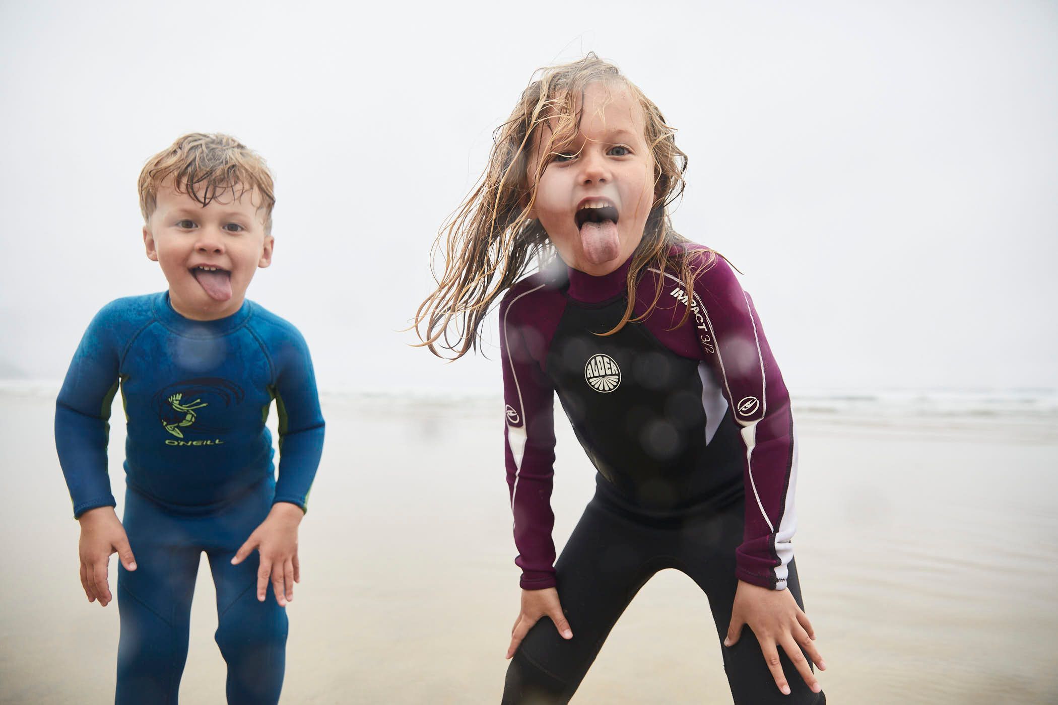 Two children in wetsuits stand on a beach, their faces covered in sand and their hair matted with salt. They are both sticking out their tongues at the camera, looking.