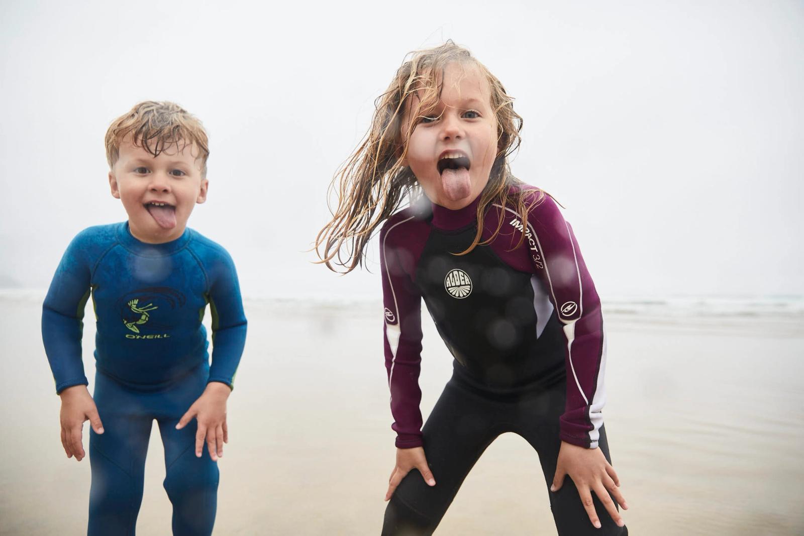 Two children in wetsuits stand on a beach, their faces covered in sand and their hair matted with salt. They are both sticking out their tongues at the camera, looking.