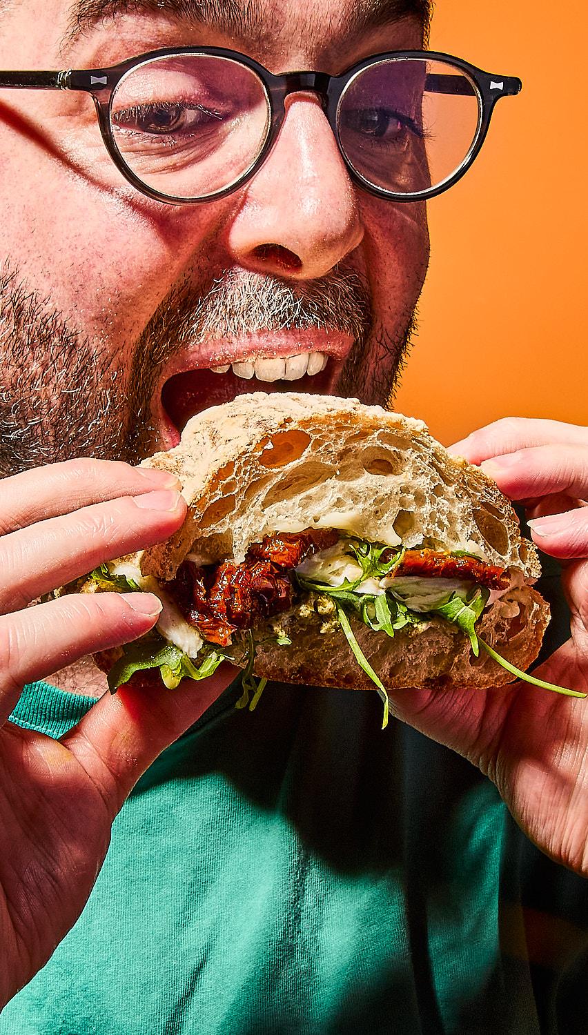 The image shows a close-up of a man with glasses and a beard about to eat a large ciabatta sandwich filled with sun-dried tomatoes and rocket. His mouth is open in anticipation against a bright orange background.