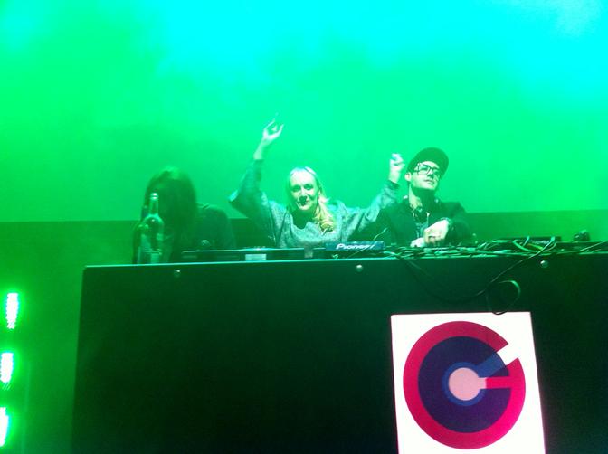A group of people, including a woman with long hair and a man with a hat, standing behind a DJ booth, surrounded by music equipment and lights.