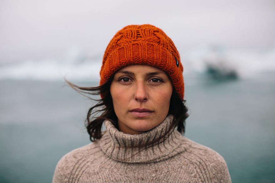 A woman wearing a red beanie and a beige turtleneck sweater, standing in front of a blurred ocean background.