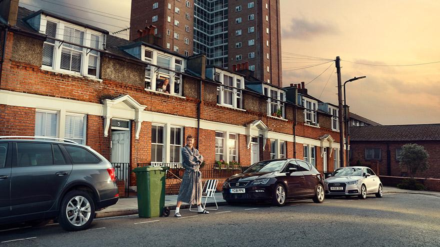 A woman in a grey dress stands in front of a row of red brick houses with white trim, with a green trash bin and a white chair nearby.