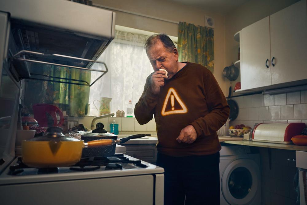 A man in a kitchen, standing near a stove, holds food to his nose and looks down at the stove, with a warning sign visible.