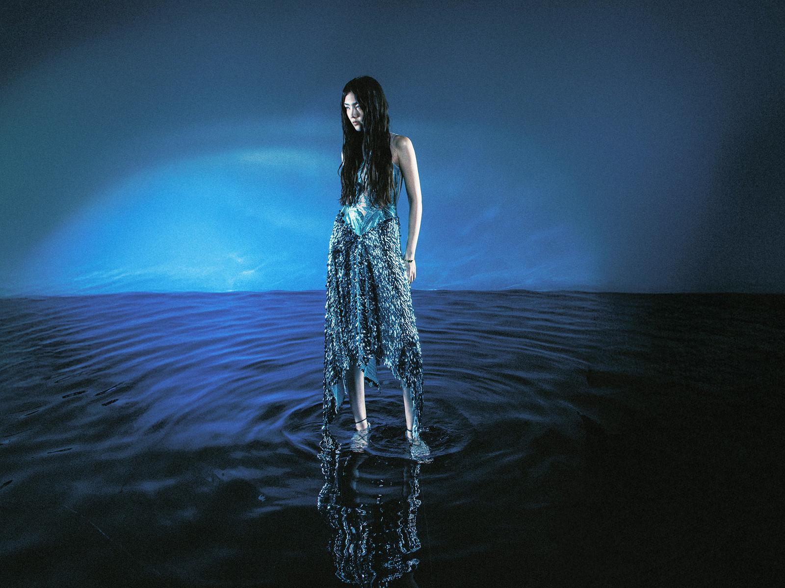 A woman stands in water, wearing a dark blue dress, with wet hair and dress.