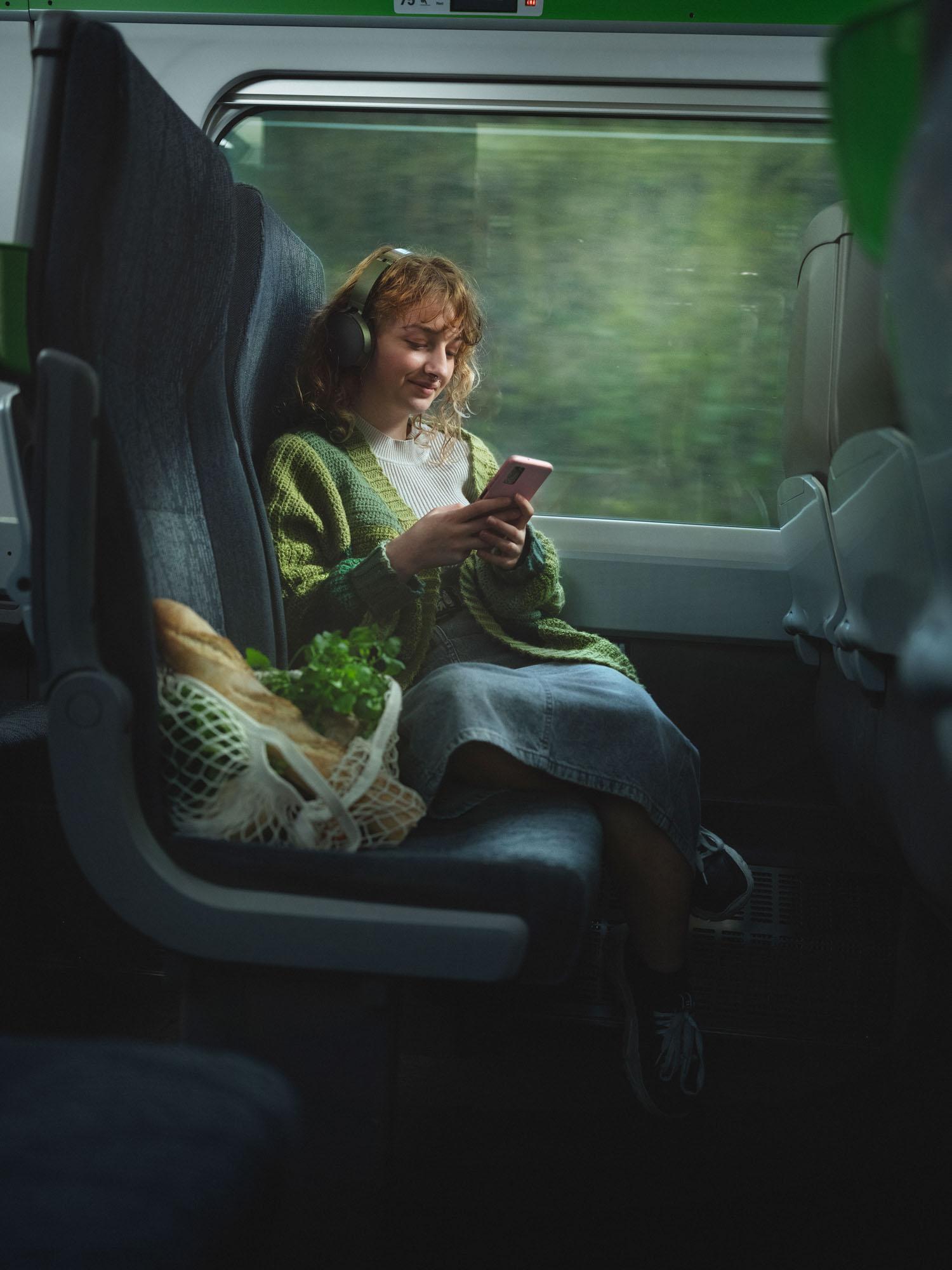 A woman with curly red hair, wearing a green cardigan, white top, and denim skirt, sitting on a train with a phone in her hand, surrounded by a shopping bag with bread and a plant.