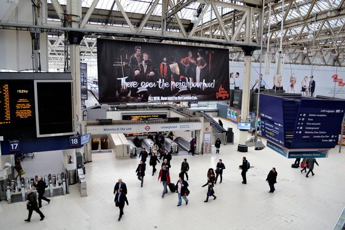 A large, empty, and dimly lit underground station with a large billboard featuring a group of people in historical costumes, with a large, red, London Dungeon logo in the top left corner.