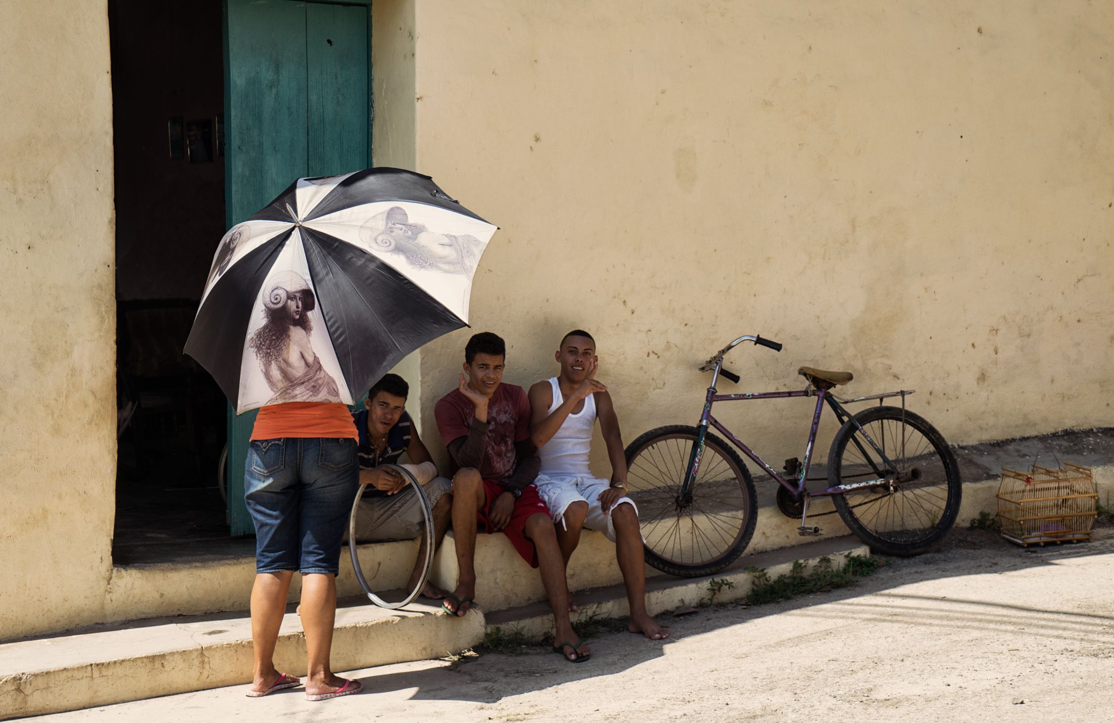 A woman holds an umbrella with a picture of a man while four men sit on steps behind her, with a bicycle and bird cage in the background.