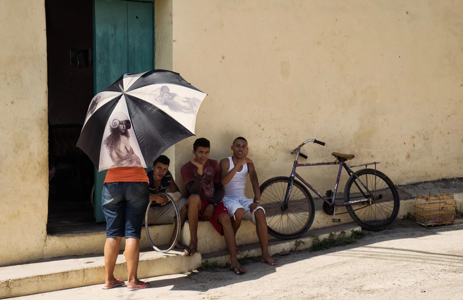 A woman holds an umbrella with a picture of a man while four men sit on steps behind her, with a bicycle and bird cage in the background.