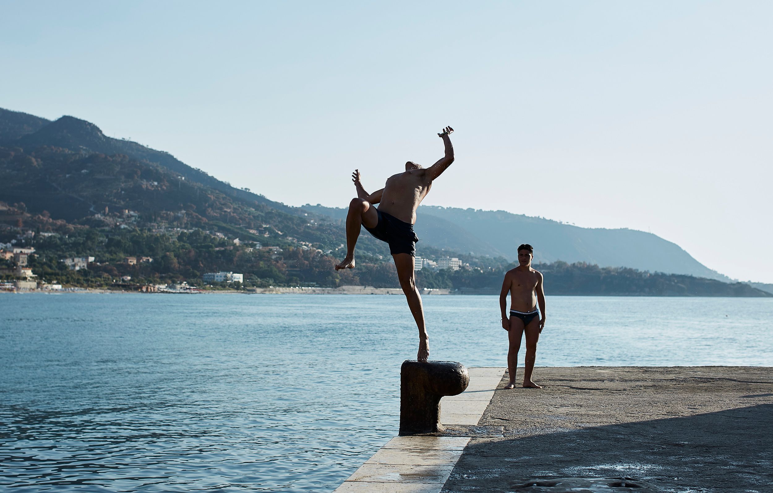 A man in black swimwear is jumping off a rock into the water, with a boy in swimwear standing on the rock, both in a scenic coastal location.