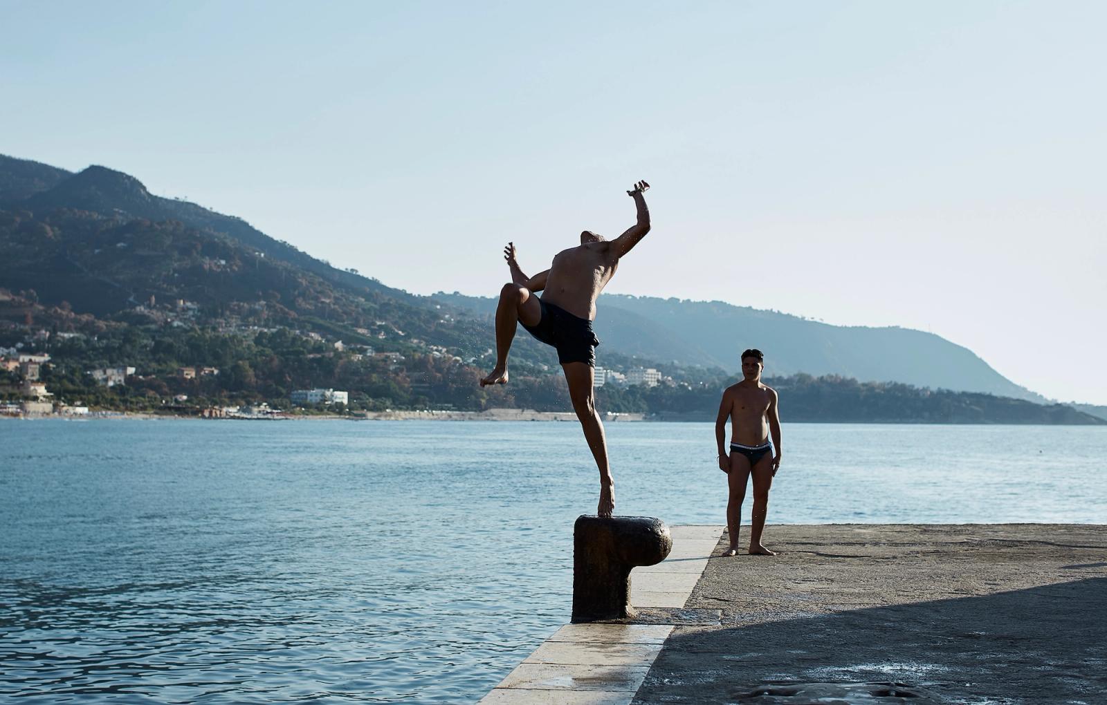 A man in black swimwear is jumping off a rock into the water, with a boy in swimwear standing on the rock, both in a scenic coastal location.