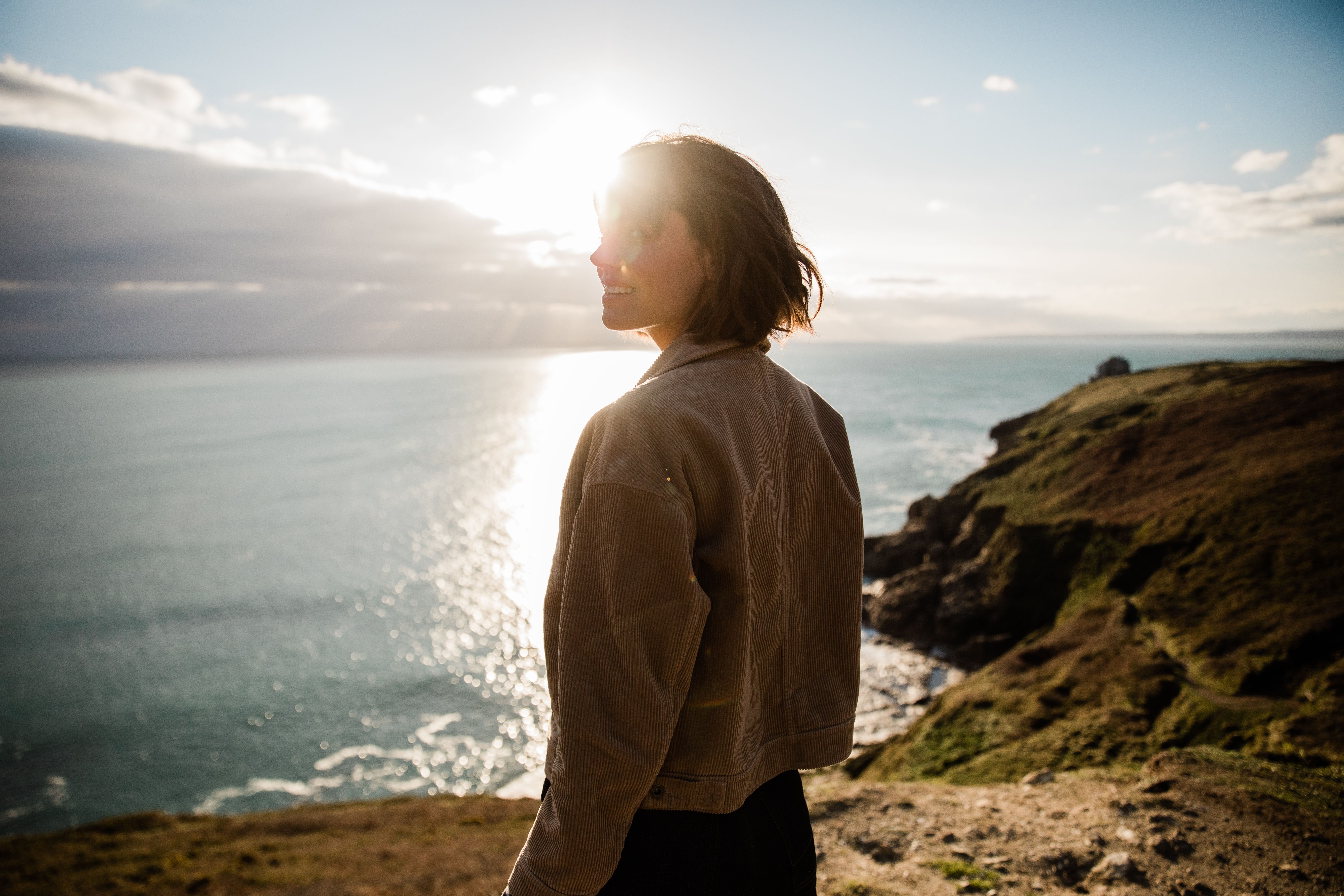 A woman with short hair and a beige jacket standing on a cliff overlooking the ocean at sunset.