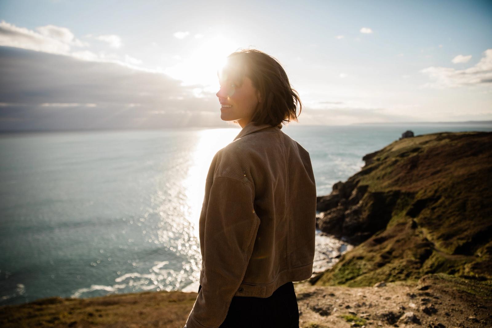 A woman with short hair and a beige jacket standing on a cliff overlooking the ocean at sunset.