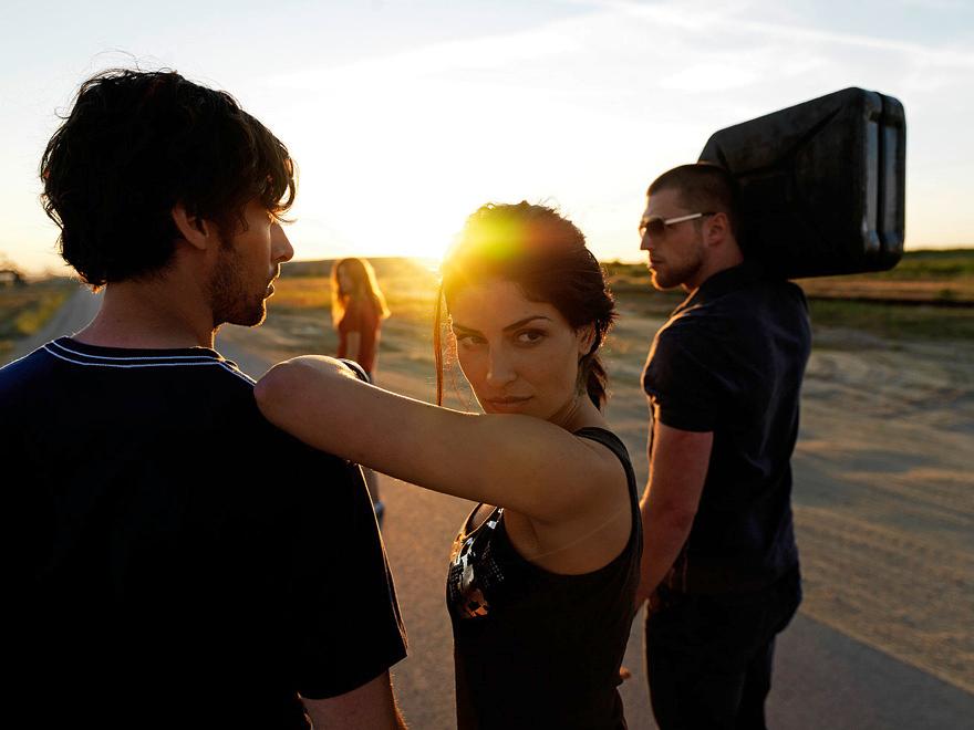 A woman in a black tank top and a man in a black t-shirt standing on a road, with a woman in the background and a man carrying a large object on his head.