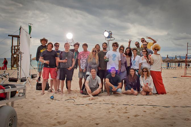 A group of people on a beach, some wearing casual clothing and others in wetsuits, posing for a photo with a camera and lighting equipment.
