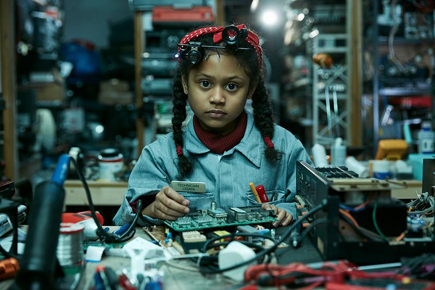 A young girl with braids and a denim jacket sits at a workbench surrounded by electronics and tools, holding a circuit board.