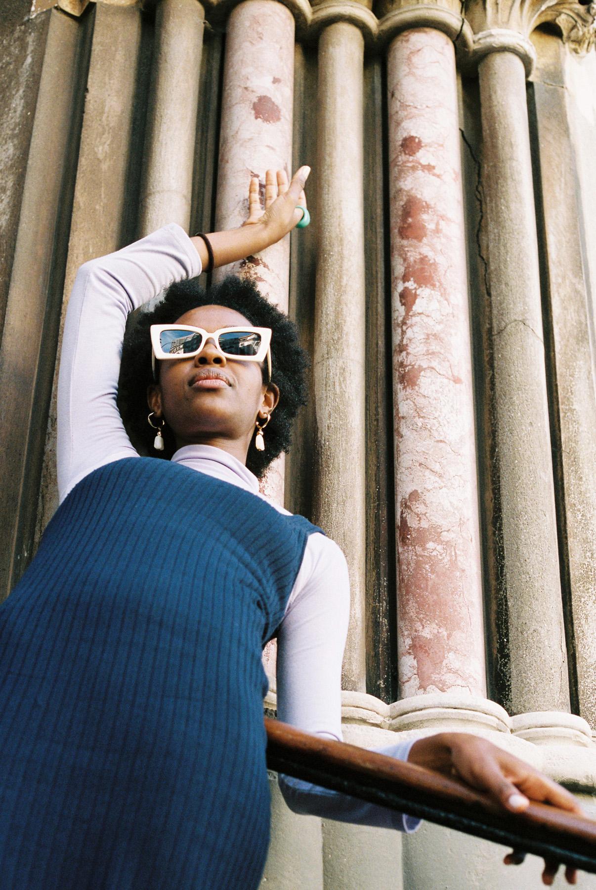 A woman with a short, curly afro and a blue sweater vest stands in front of a stone wall with ornate columns, looking up at the camera with a confident expression.