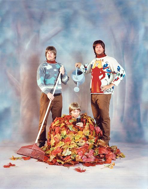 Two men and a child in a forest, one man holding a broom and the other a watering can, surrounded by a large pile of autumn leaves.