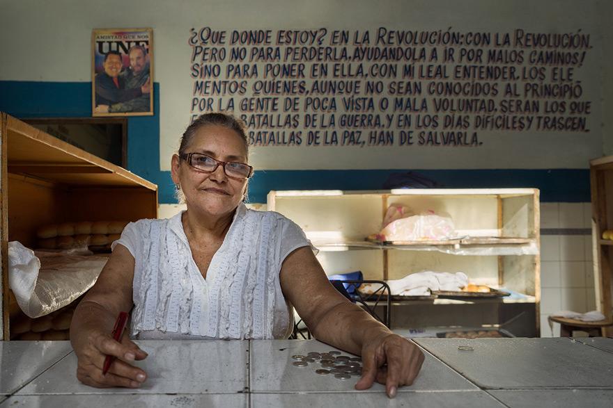 A woman in a white blouse sits at a counter in a bakery or similar food service environment, with a wall behind her containing a large piece of writing in a foreign language.