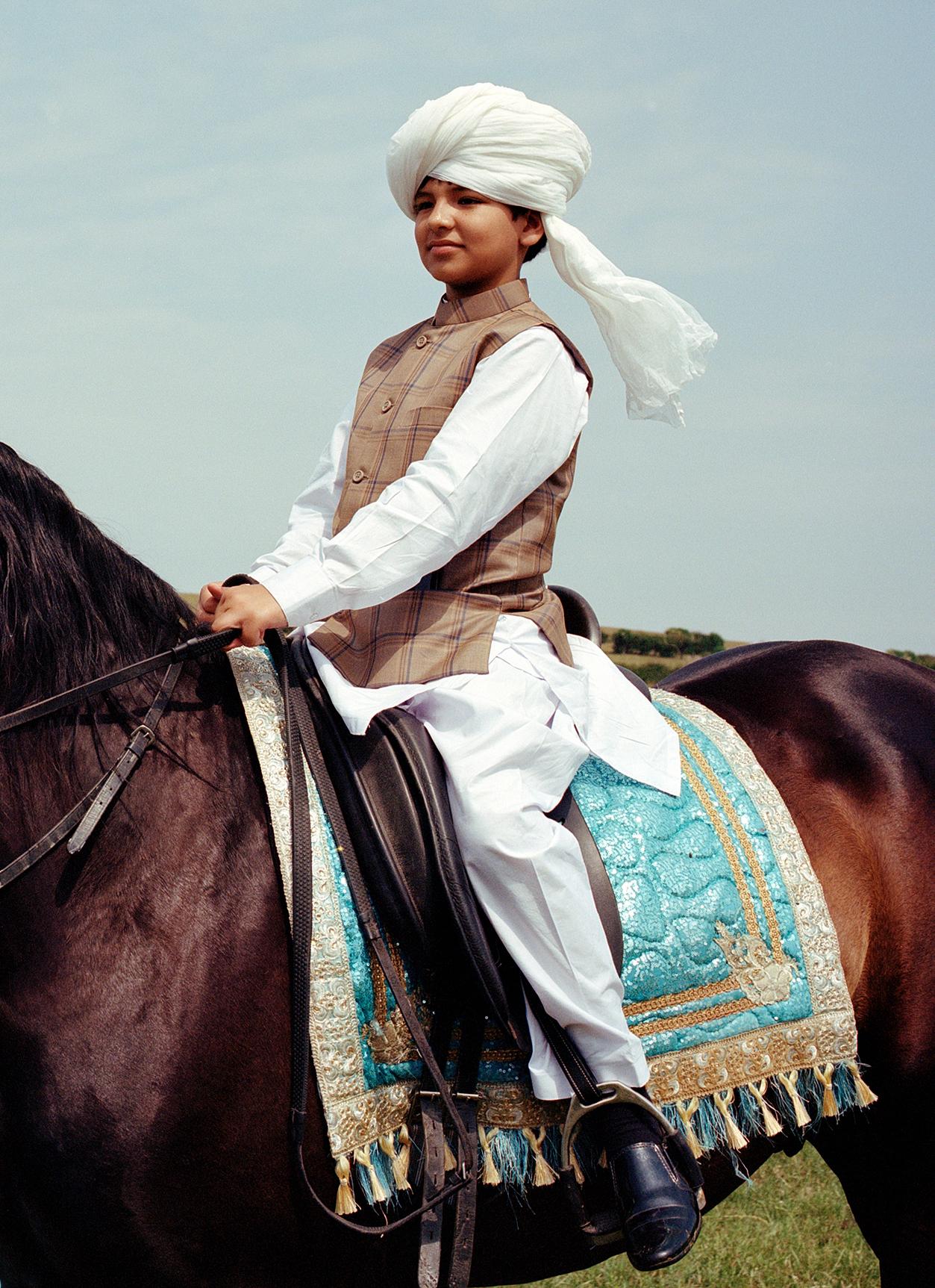 A young boy in a white turban and traditional Pakistani clothing riding a brown horse in a grassy field.