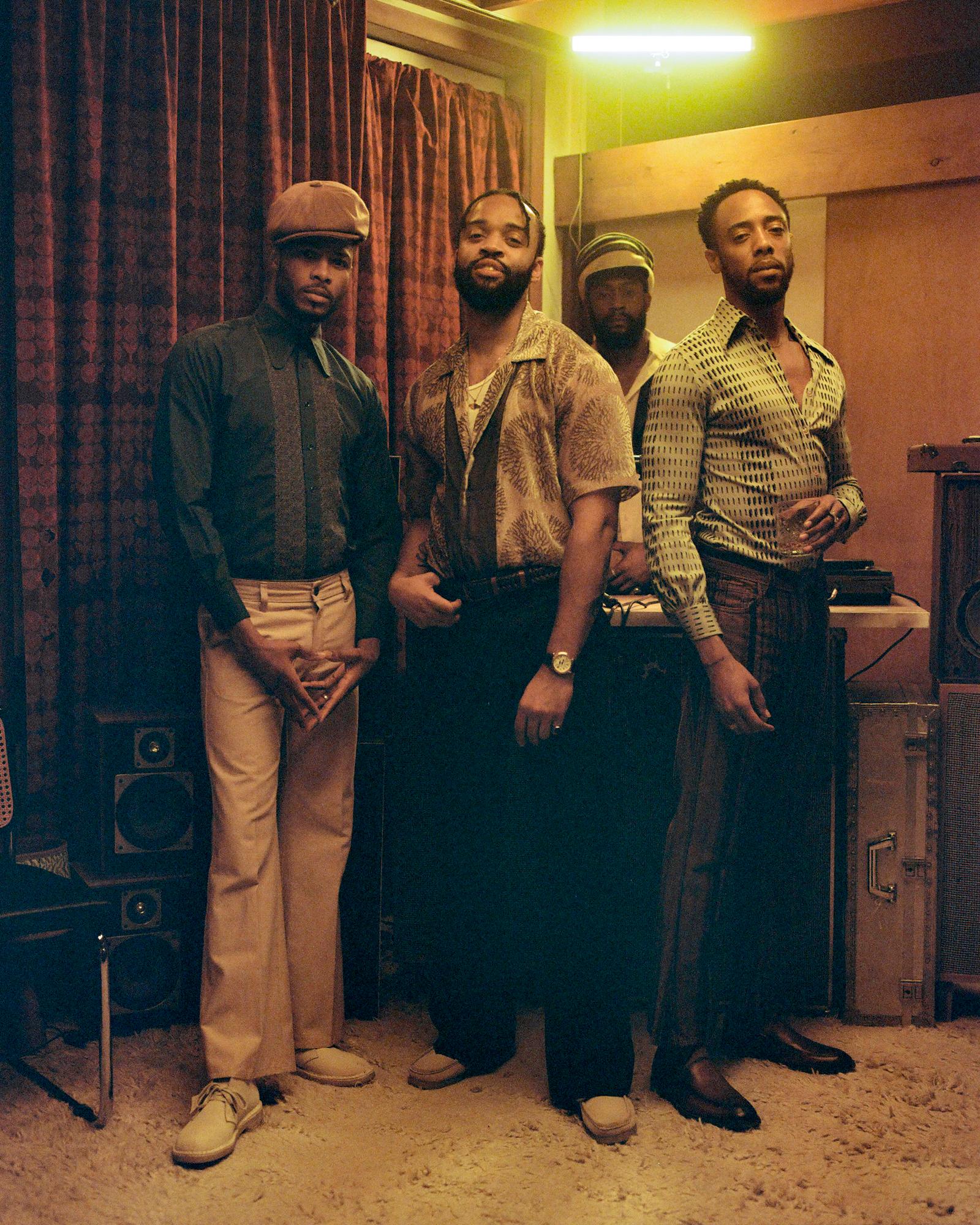 Four men in 1970s clothing stand in a dim room with vintage and modern equipment.