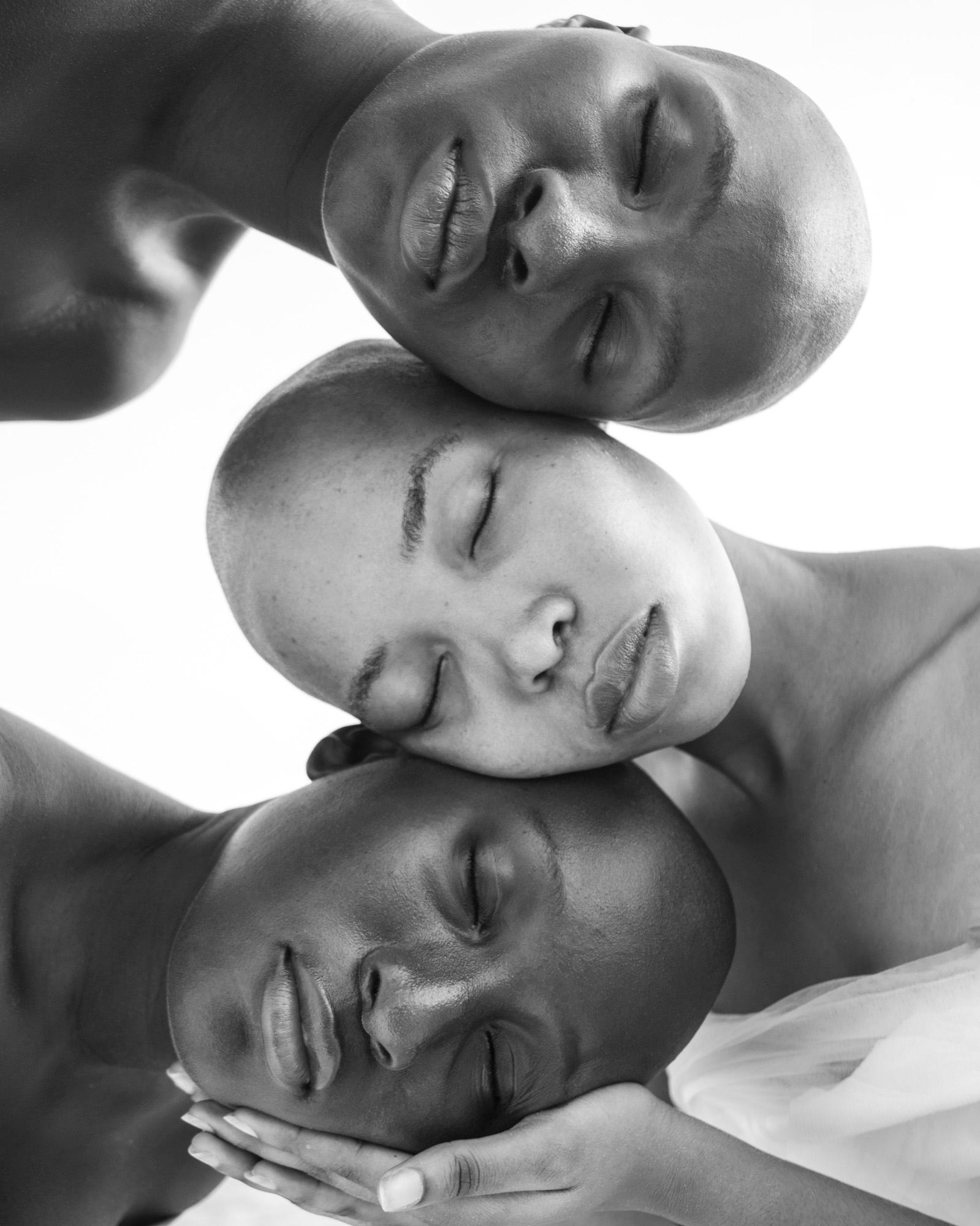 Three people of different skin tones and hair textures, all with their eyes closed, resting their heads on top of each other, creating a human pyramid.