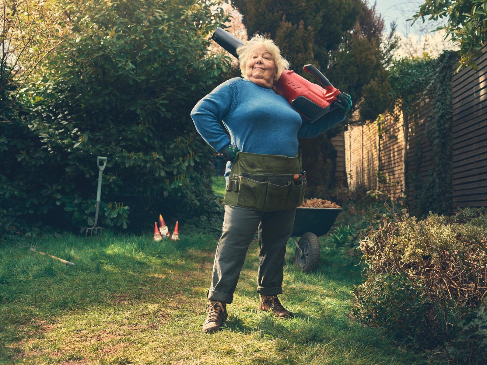 A woman in her 60s stands in a garden with a lawnmower on her shoulder, surrounded by gardening tools and a wheelbarrow.