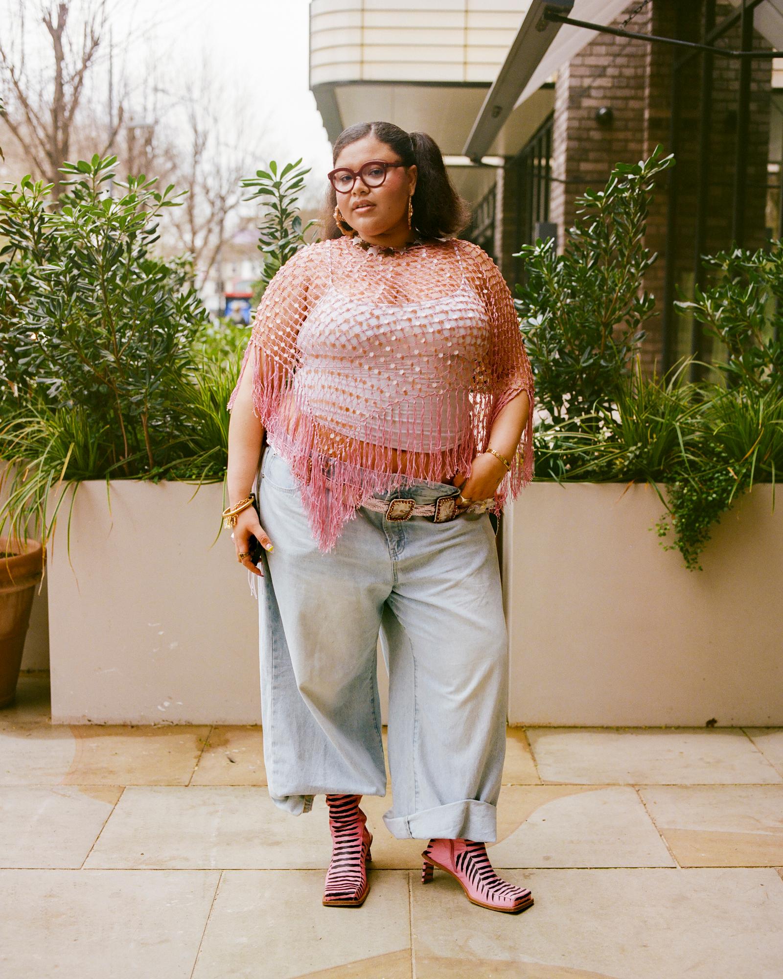 A woman wearing a pink sequined top, light blue jeans, and pink and black striped high-heeled ankle boots, standing in front of a white wall with green plants.