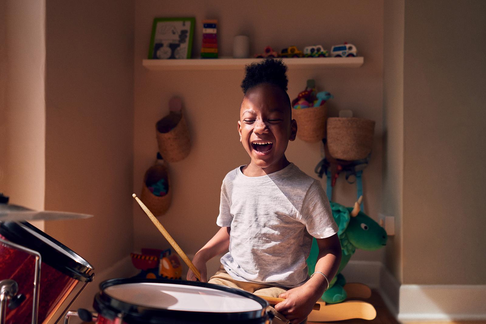 A young boy with a big smile is playing a drum set in a room filled with toys and music equipment.