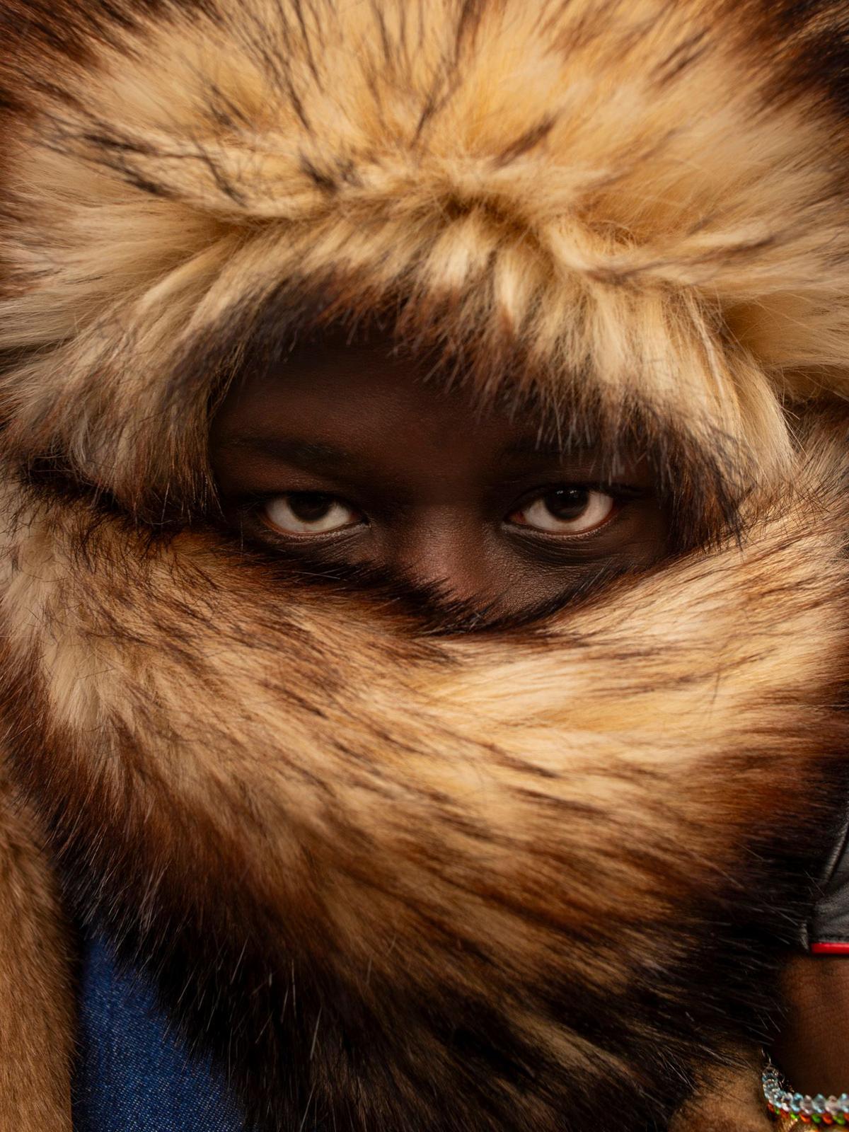 A person wearing a fur-lined coat with a distinctive, oversized, and fluffy fur-trimmed hood, with a visible face and eyes looking directly at the camera.