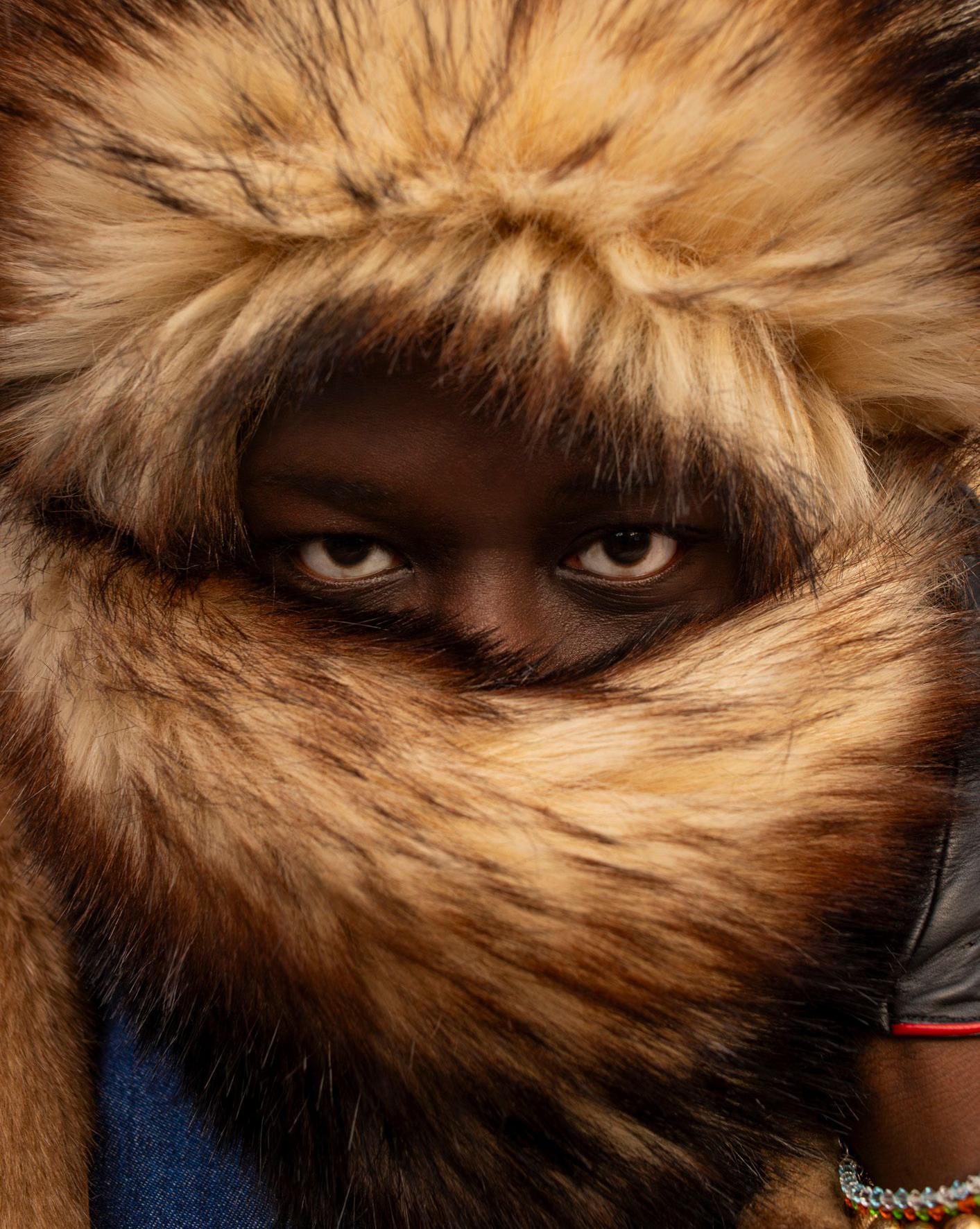 A person wearing a fur-lined coat with a distinctive, oversized, and fluffy fur-trimmed hood, with a visible face and eyes looking directly at the camera.