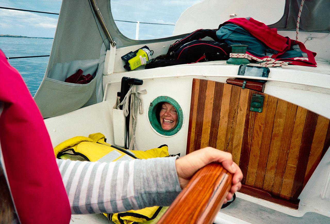 A woman is peeking out of a porthole on a boat, wearing a grey and white striped long-sleeve shirt.