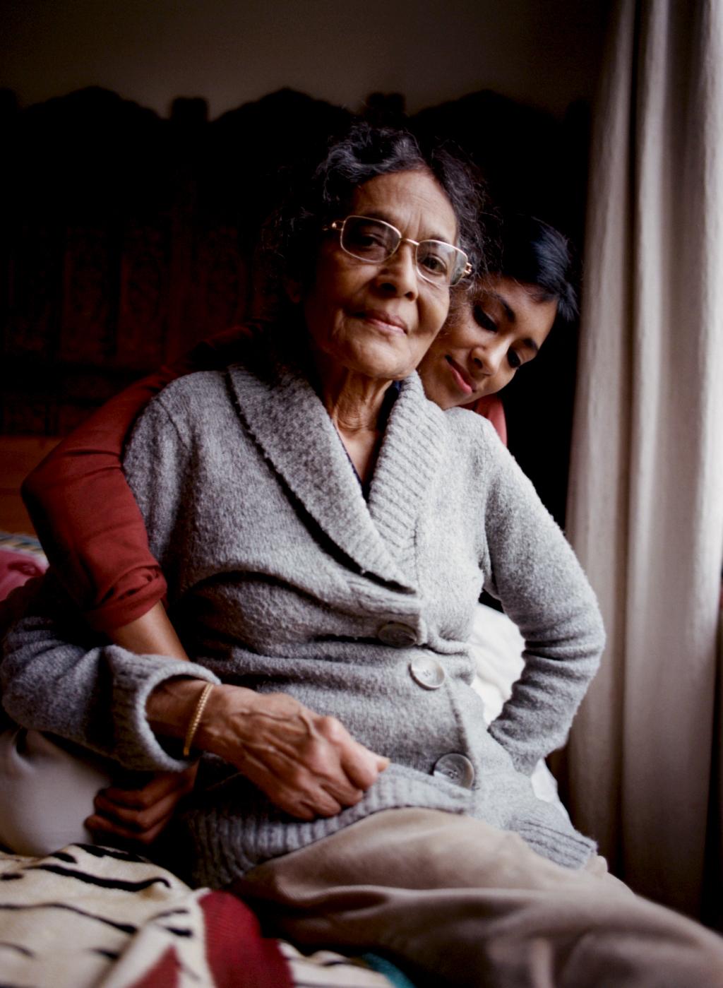 Two women with dark hair sit on a bed, one older and wearing glasses, the other younger and in a red shirt, both looking at the camera seriously.